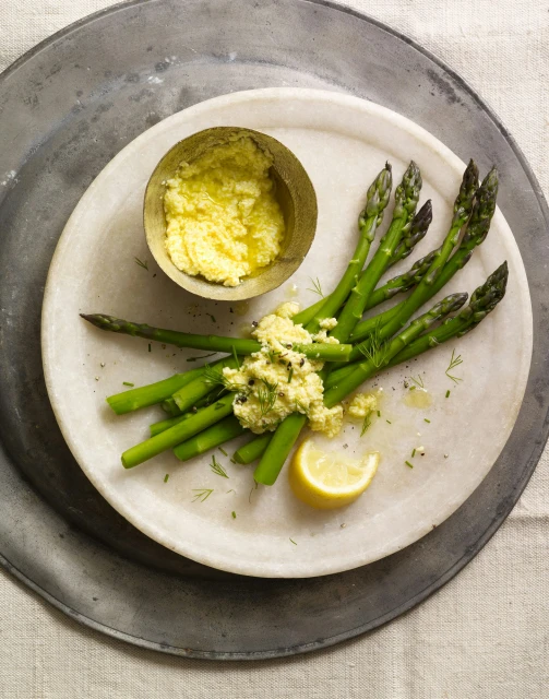 Overhead shot of asparagus spears topped with sauce, served with a lemon wedge on a marble plate and rustic metal surface.