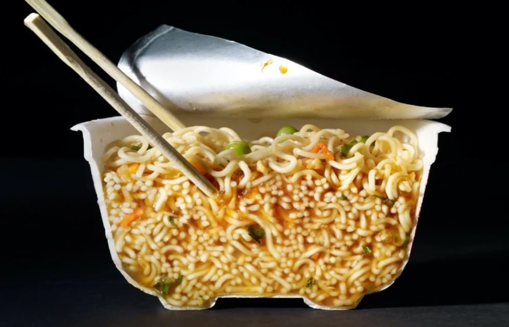 Overhead shot of open cup of instant ramen noodles with chop sticks, featuring noodles, broth, peas, and carrots against dark background.