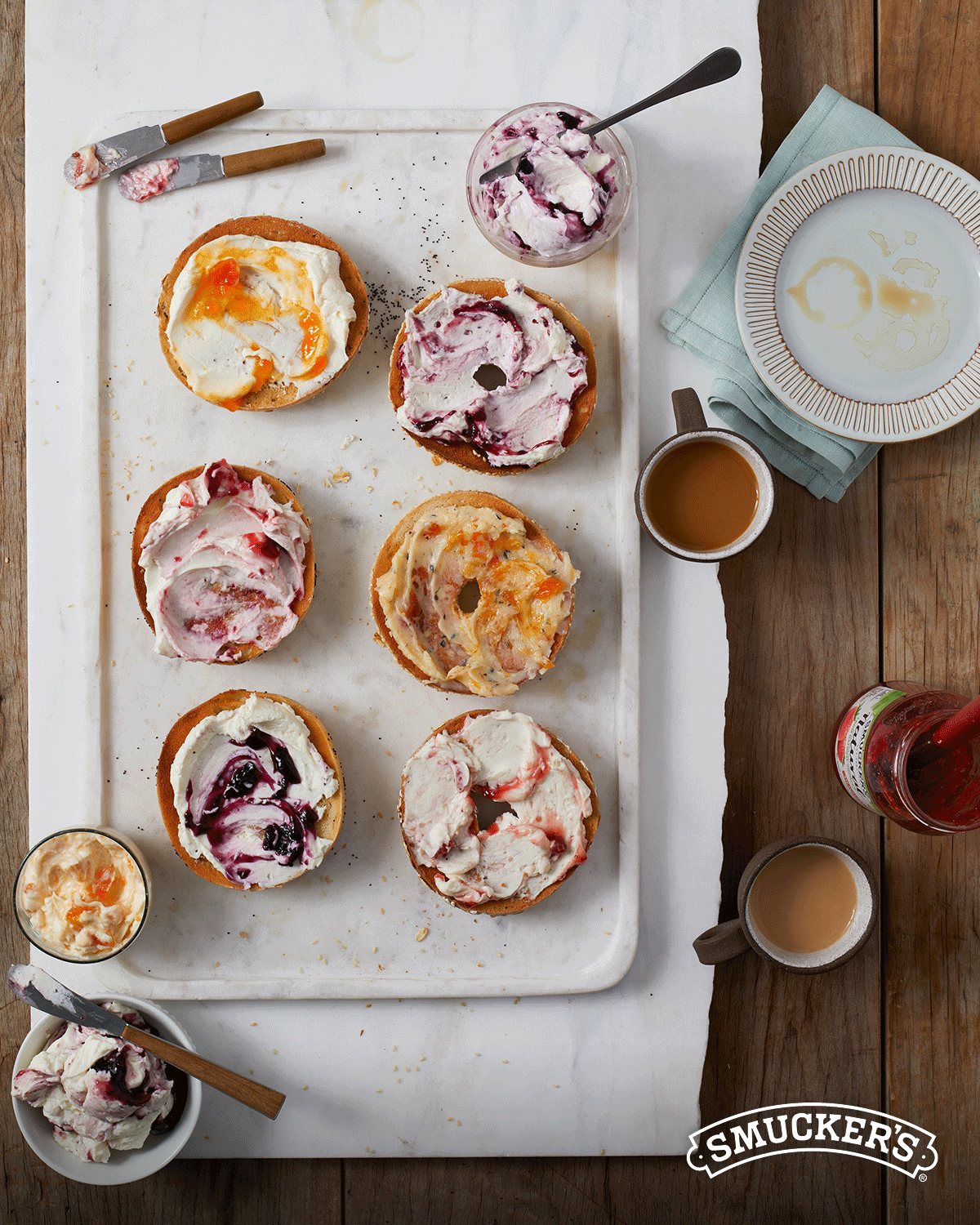 Overhead shot of bagels topped with cream cheese and jam, served with coffee and jam jars on a white tray atop a wood surface.