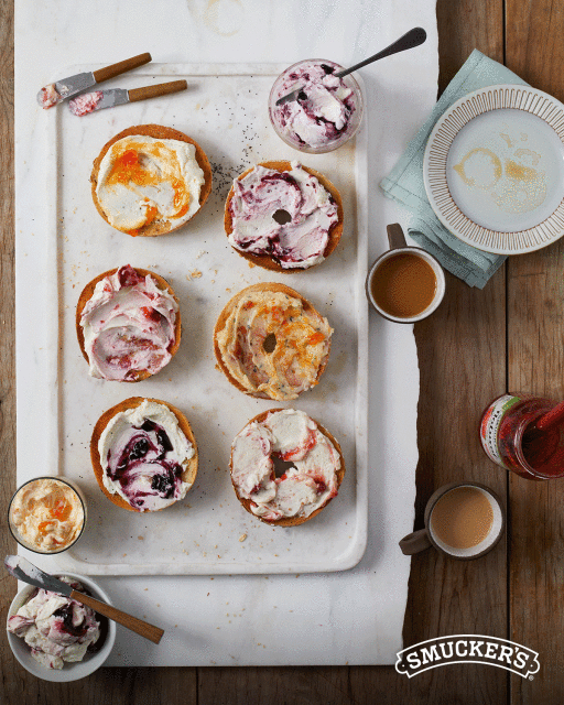 Overhead shot of bagels topped with cream cheese and jam, served with coffee and jam jars on a white tray atop a wood surface.