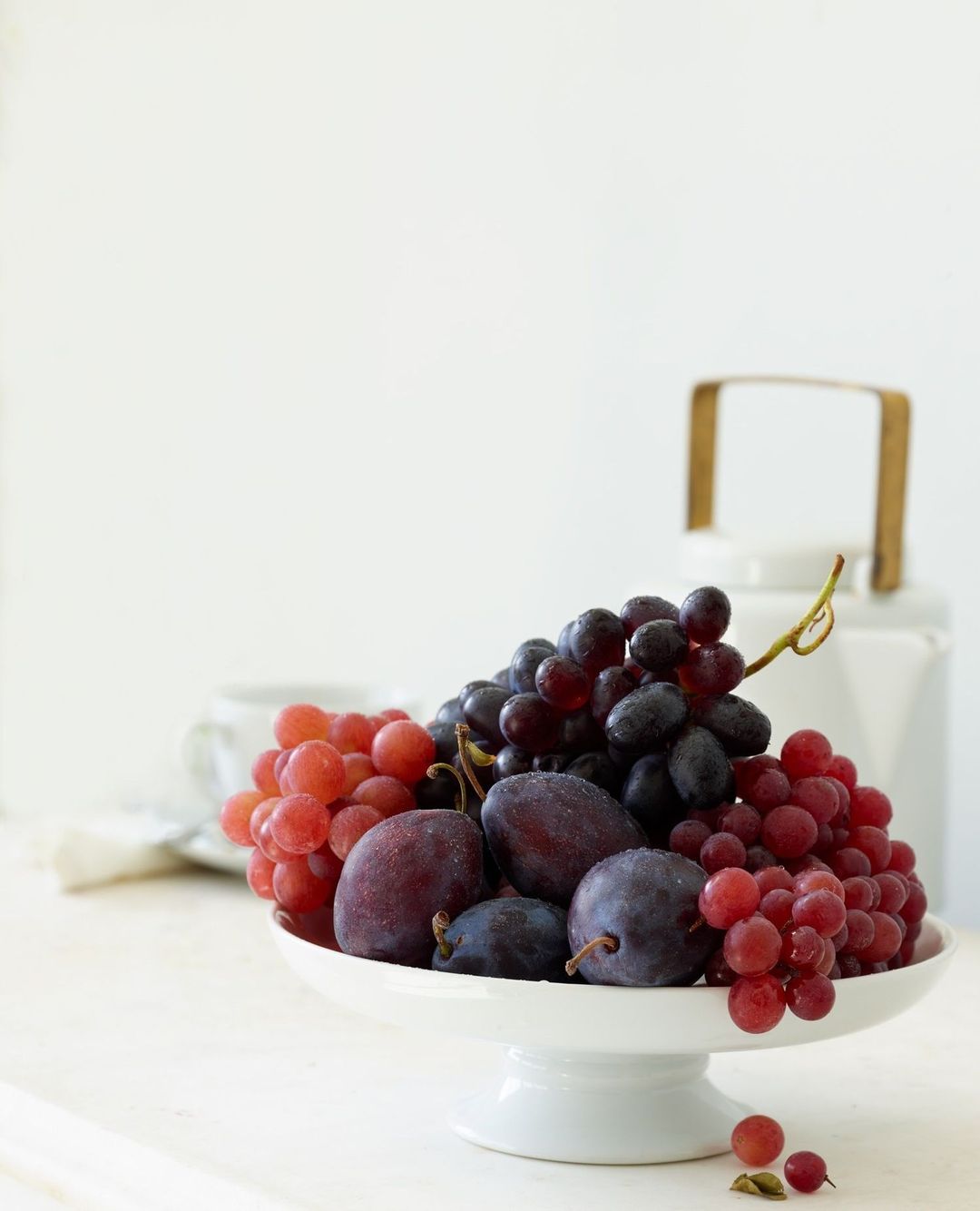 Still life of red and purple grapes, and plums on a white pedestal cake stand with a white teapot and teacup in the background.