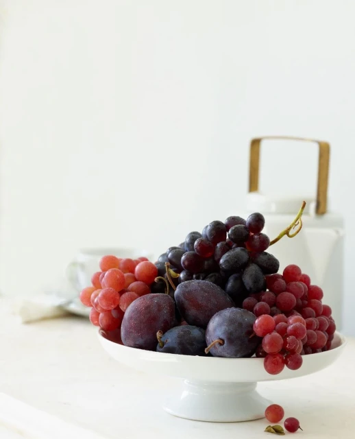 Still life of red and purple grapes, and plums on a white pedestal cake stand with a white teapot and teacup in the background.