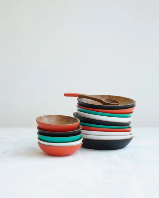 Stacks of colorful wooden bowls and a small wooden spoon are arranged on a white marble surface against a white background.