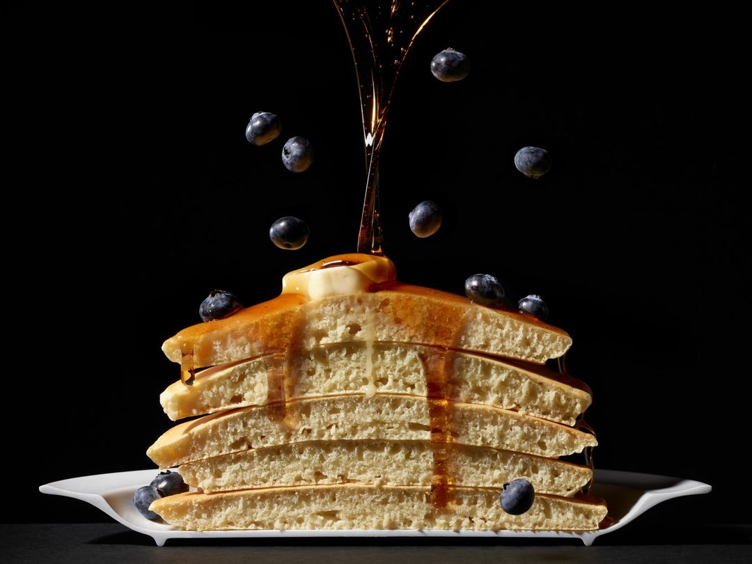 Stack of pancakes on a white plate with maple syrup pouring over them and blueberries scattered on the pancakes and around them.