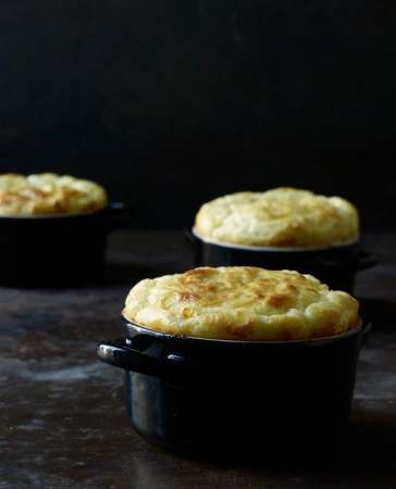 Three souffle desserts baked in small black ramekins sit on a dark surface against a dark background in a moody food photography shot.