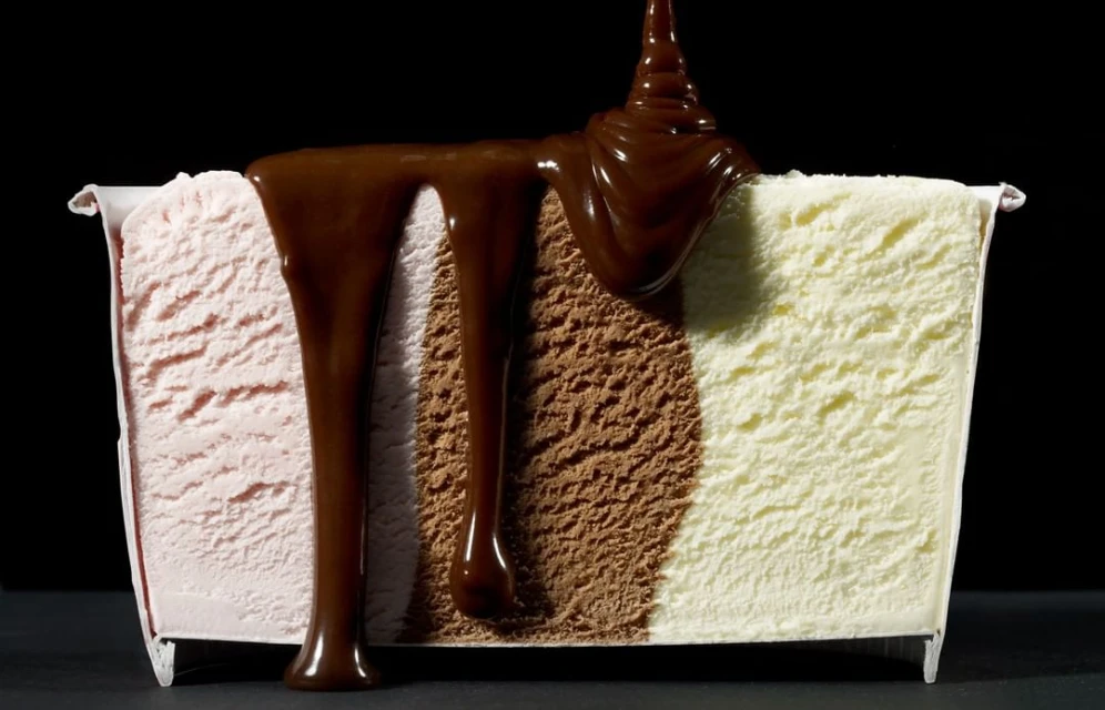 Close-up of a cut container of Neapolitan ice cream with vanilla, chocolate, and strawberry flavors being drizzled with chocolate sauce.