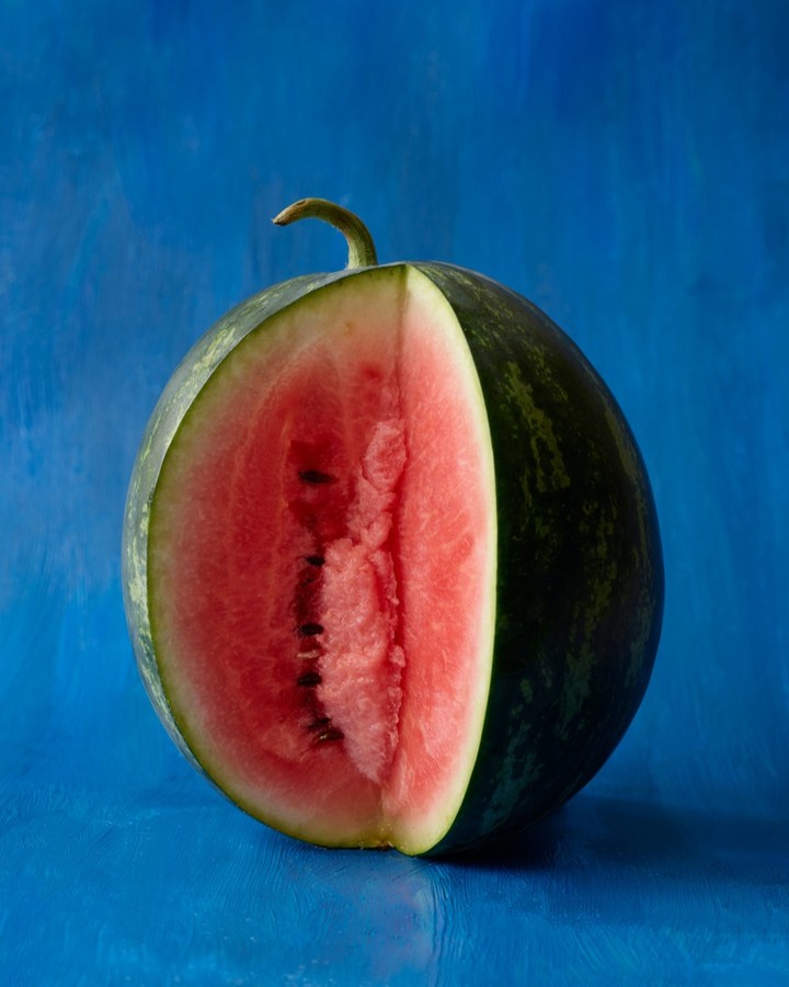 A watermelon, partially cut to show the red flesh and seeds, is set against a mottled blue background.