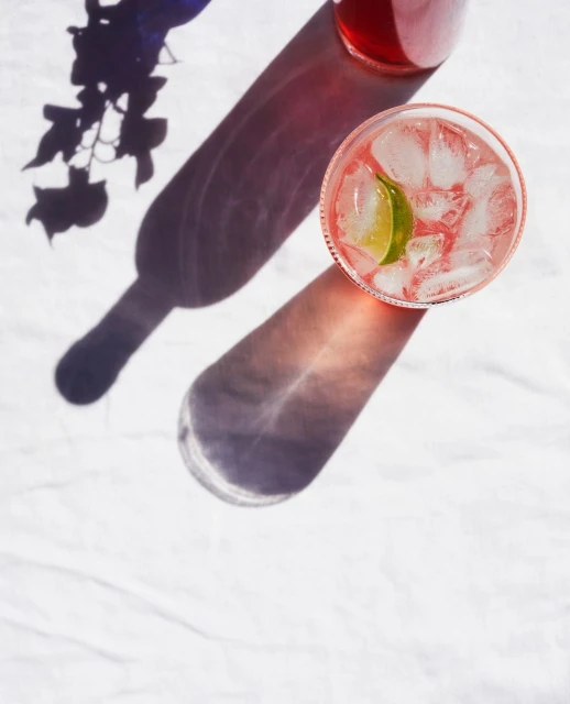 Overhead shot of a red drink with ice and lime in a glass next to a bottle, casting shadows on a white textured surface.