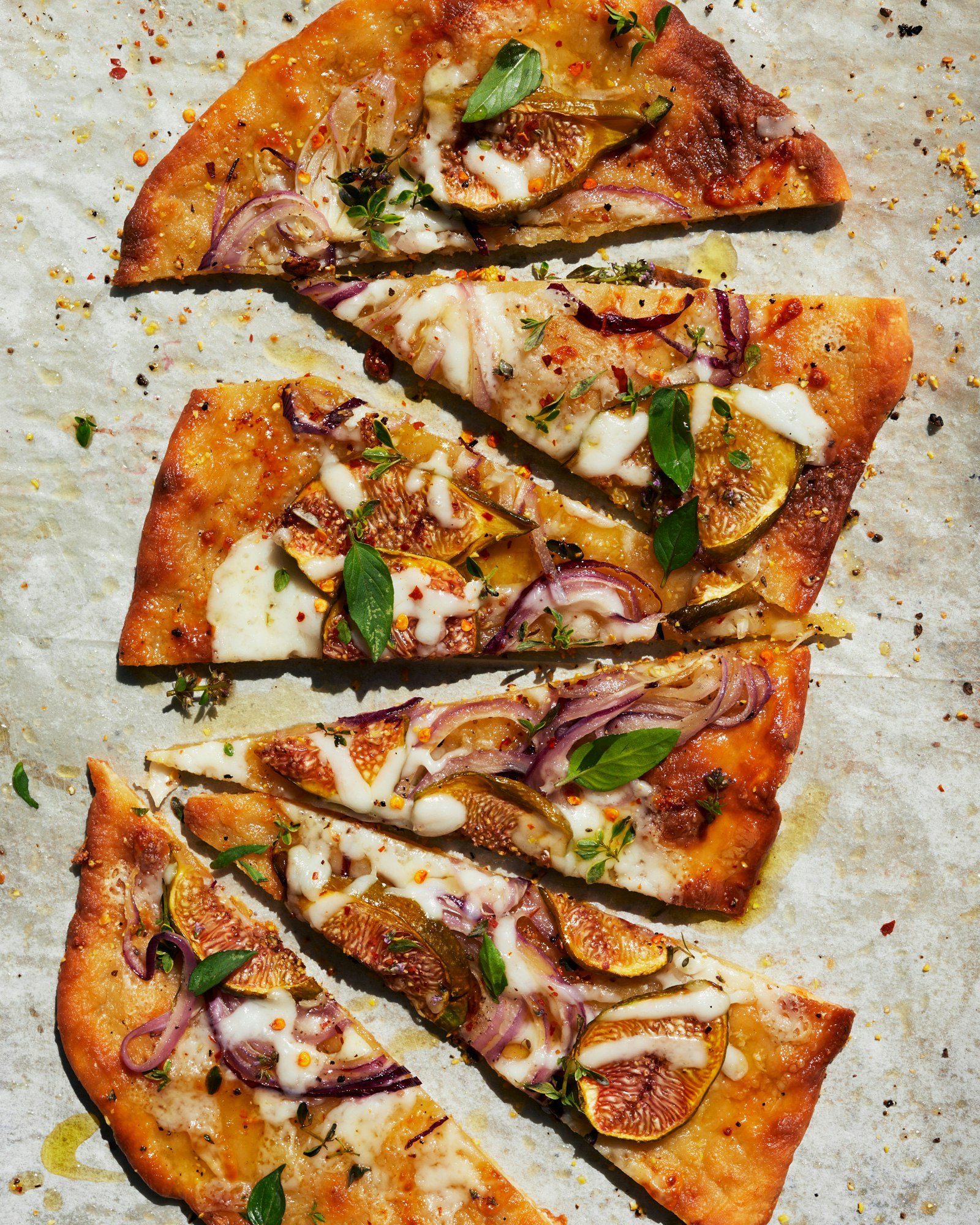 Overhead shot of a flatbread pizza cut into slices, topped with figs, red onions, basil, cheese, and spices, presented on parchment paper.