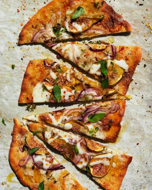 Overhead shot of a flatbread pizza cut into slices, topped with figs, red onions, basil, cheese, and spices, presented on parchment paper.