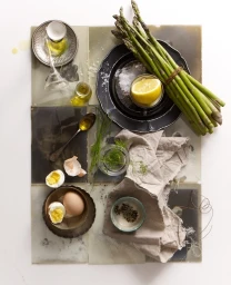 Top-down view of fresh asparagus, halved boiled eggs, lemon, olive oil, and herbs arranged on a rustic tiled surface.