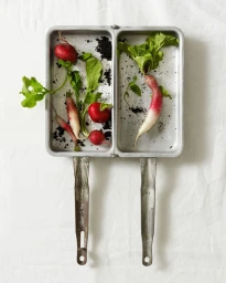 Top-down view of fresh radishes with green leaves and soil inside a dual compartment metal pan on a white cloth.