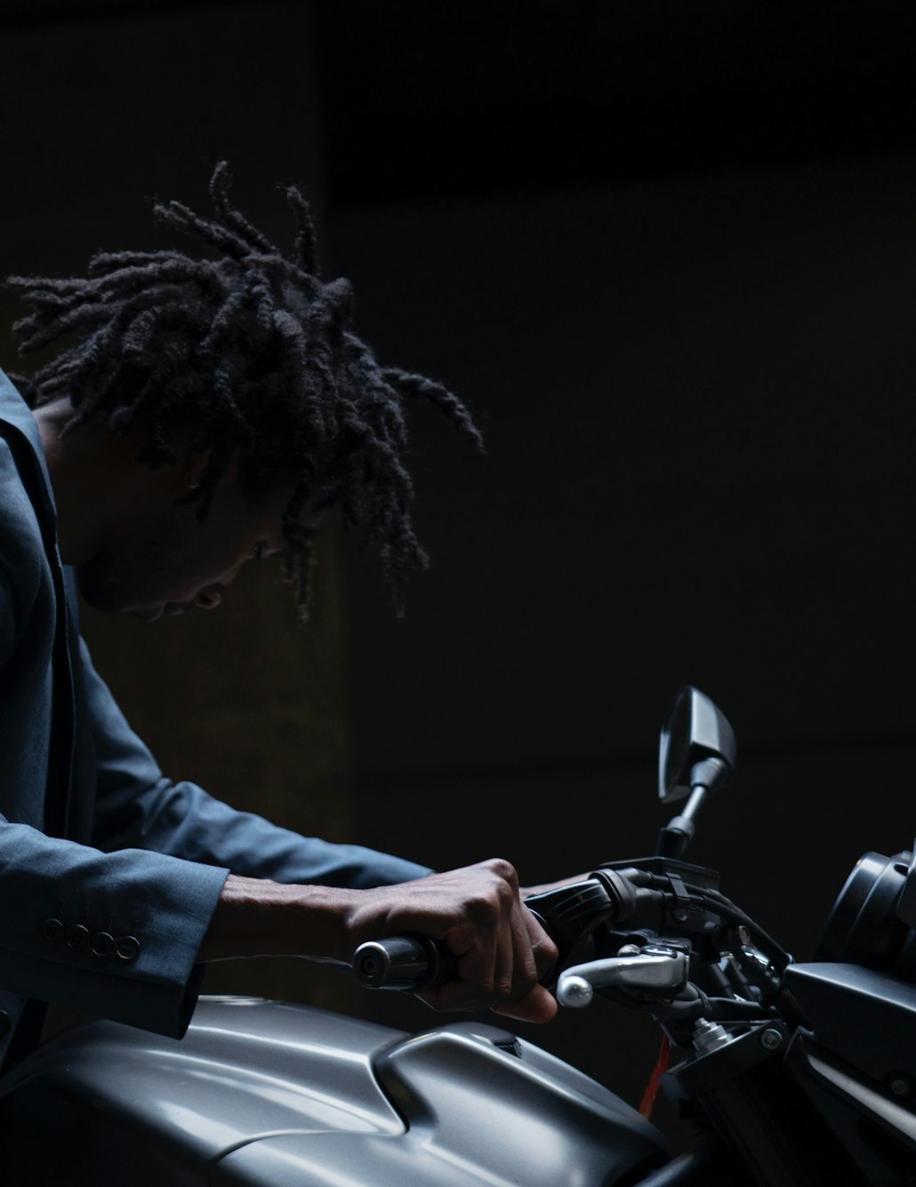 Adult male with dreadlocks in a blue suit jacket on a motorcycle with dark environment and cool lighting, conveying a relaxed mood.