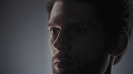 Portrait of an adult male with short brown hair and stubble, looking off-camera with thoughtful expression. Dark and moody lighting.