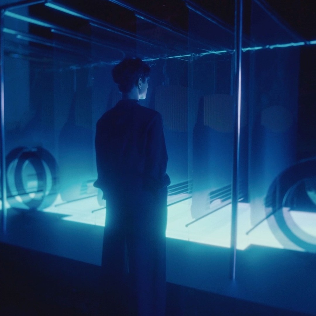 Person standing in a blue illuminated indoor space with geometric shapes. The lighting creates a futuristic, almost sci-fi atmosphere.