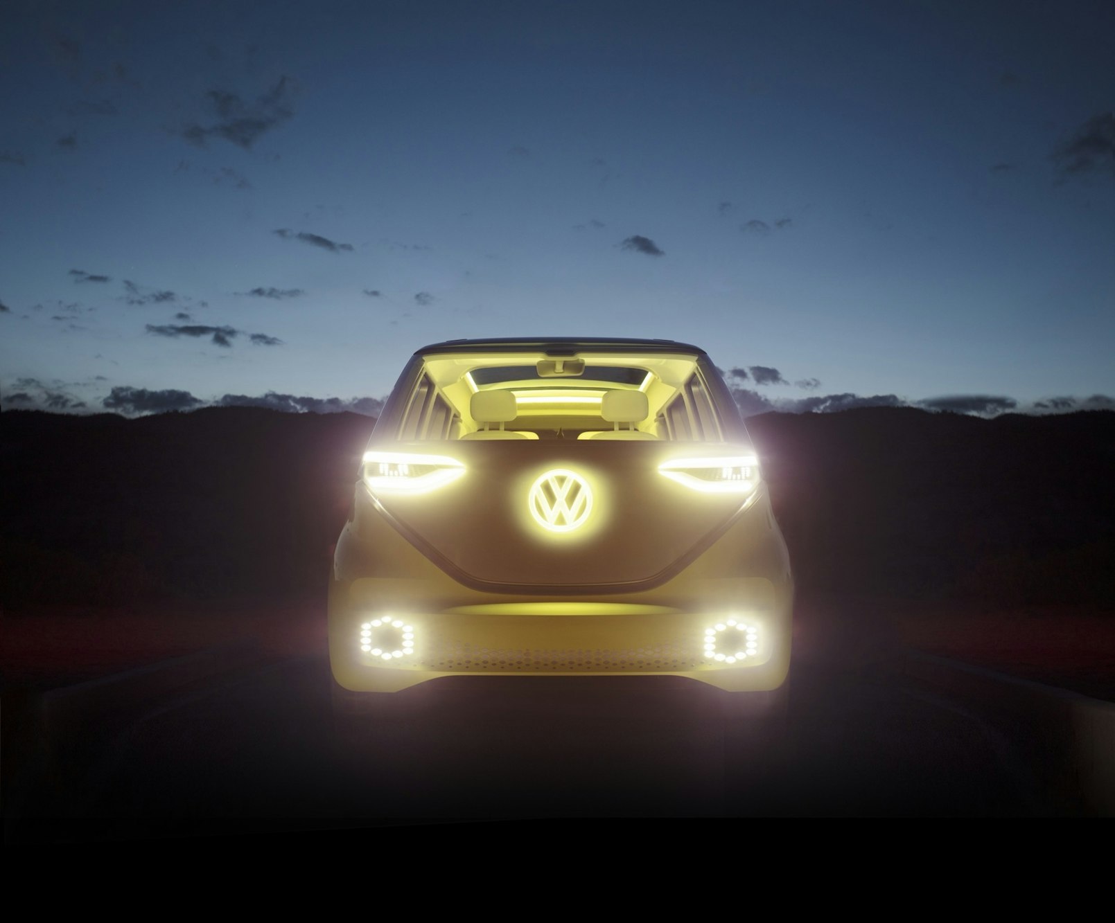 Volkswagen ID Buzz electric vehicle driving on a road at dusk, with illuminated headlights and logo, set against a dark landscape.