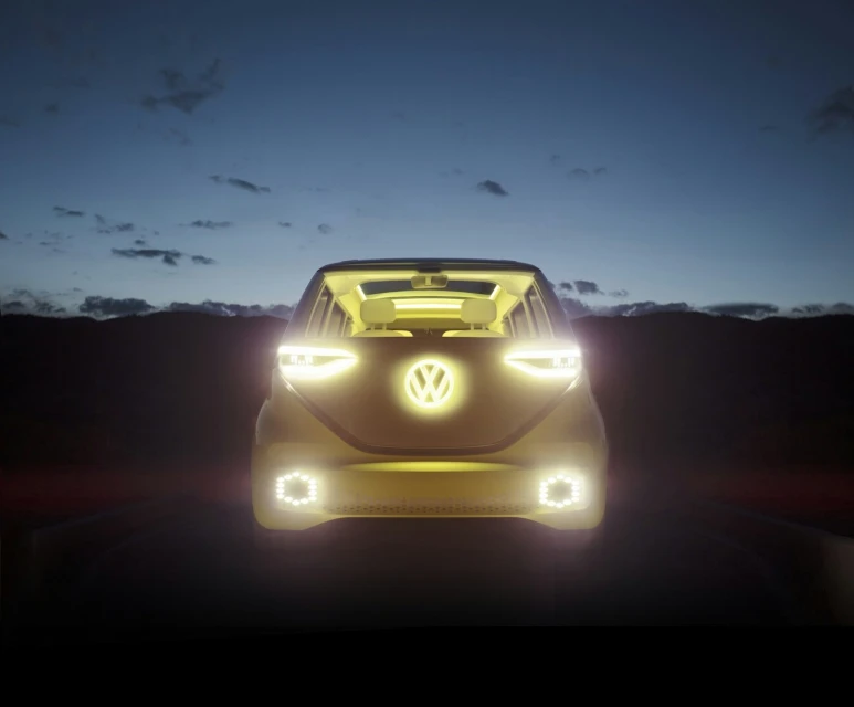 Volkswagen ID Buzz electric vehicle driving on a road at dusk, with illuminated headlights and logo, set against a dark landscape.
