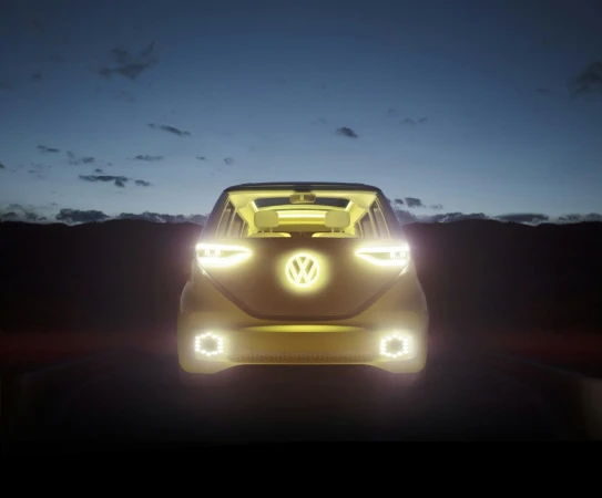 Volkswagen ID Buzz electric vehicle driving on a road at dusk, with illuminated headlights and logo, set against a dark landscape.