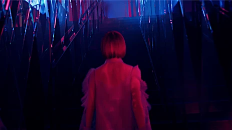From behind, a woman with a short red bob walks toward a hallway with hanging metallic streamers, lit with purple and blue lights.