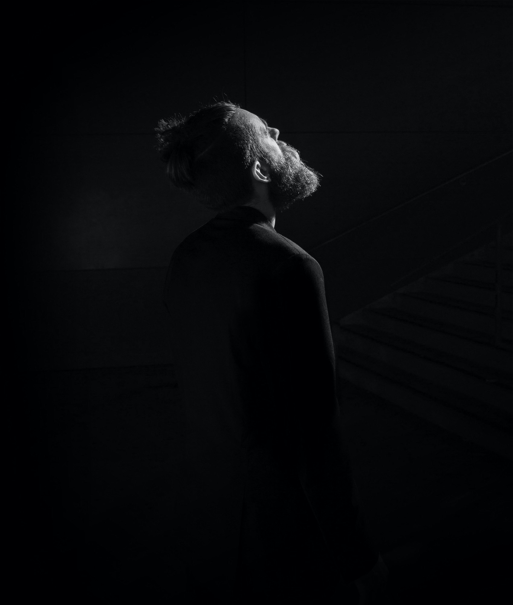 Monochrome portrait of a bearded adult male looking up in a dark setting with subtle lighting, showcasing a somber and contemplative mood.