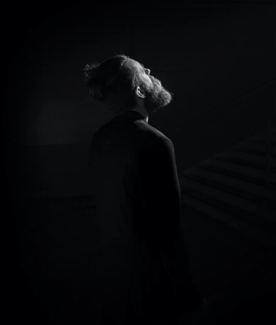 Monochrome portrait of a bearded adult male looking up in a dark setting with subtle lighting, showcasing a somber and contemplative mood.