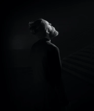 Monochrome portrait of a bearded adult male looking up in a dark setting with subtle lighting, showcasing a somber and contemplative mood.