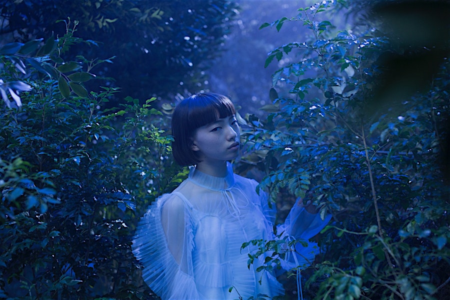Young woman in a sheer ruffled dress stands amidst lush greenery in a moody outdoor setting with a cool blue color grading.