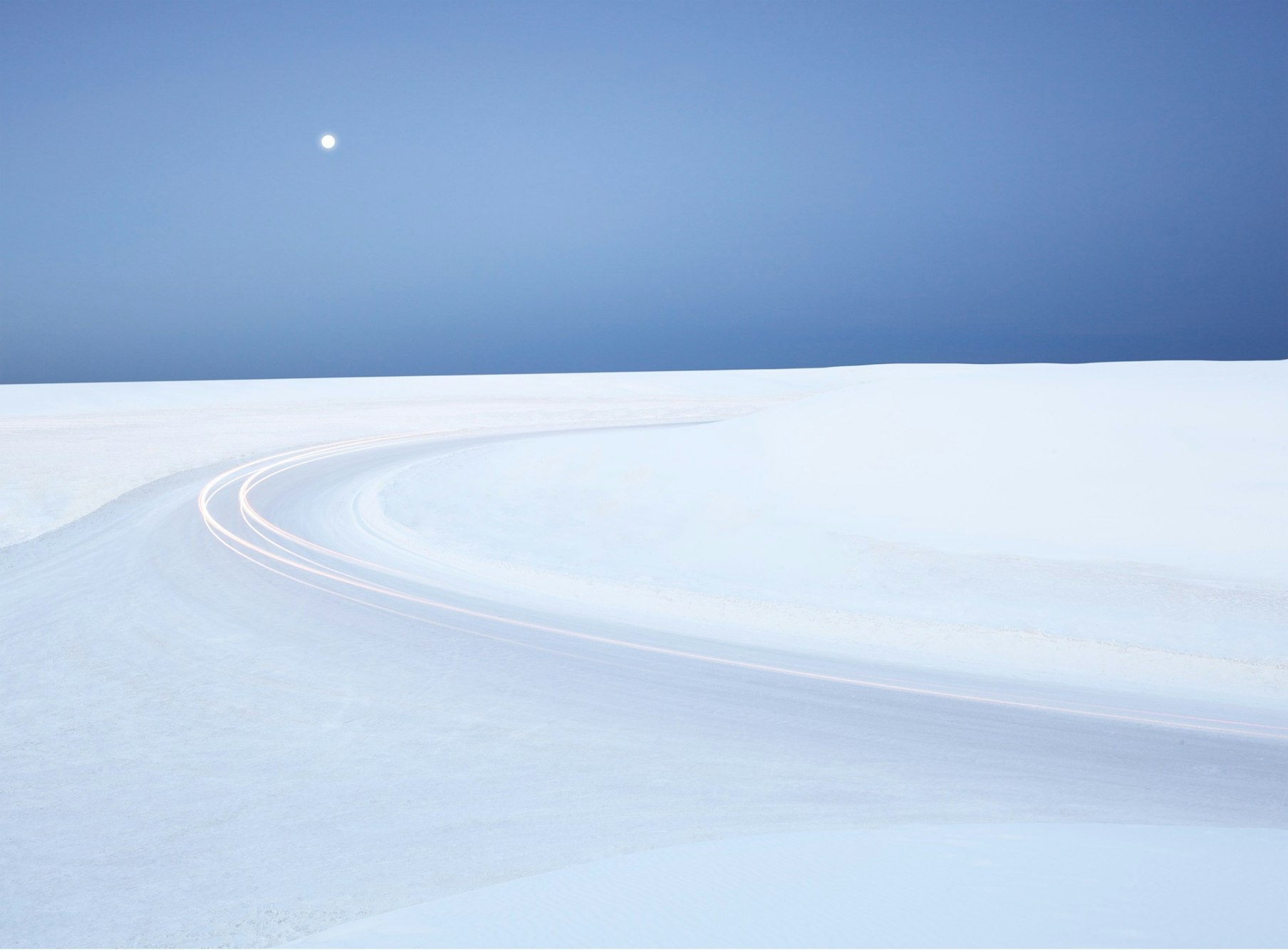 Snowy landscape with curving road and moonlight. Long exposure photography of light trails in a serene, minimal outdoor setting.