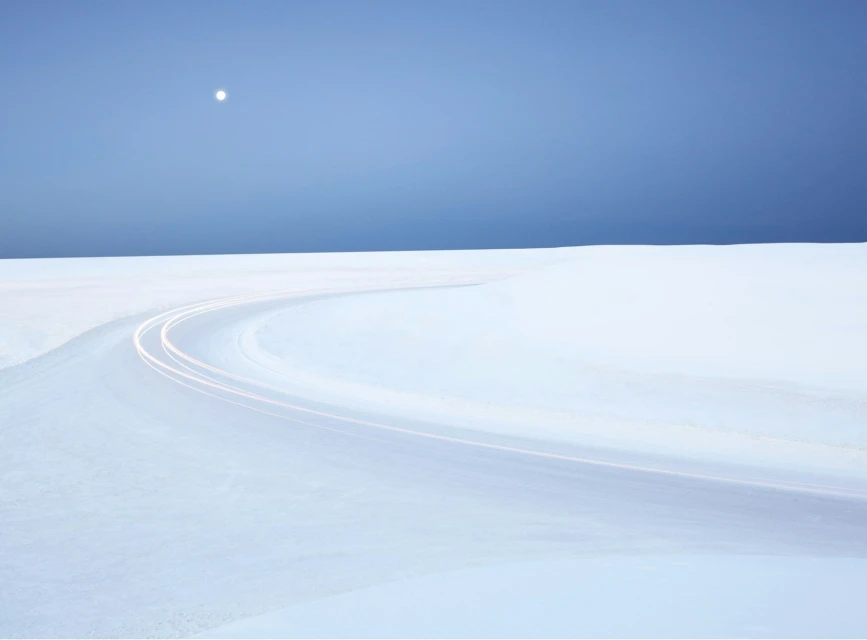 Snowy landscape with curving road and moonlight. Long exposure photography of light trails in a serene, minimal outdoor setting.
