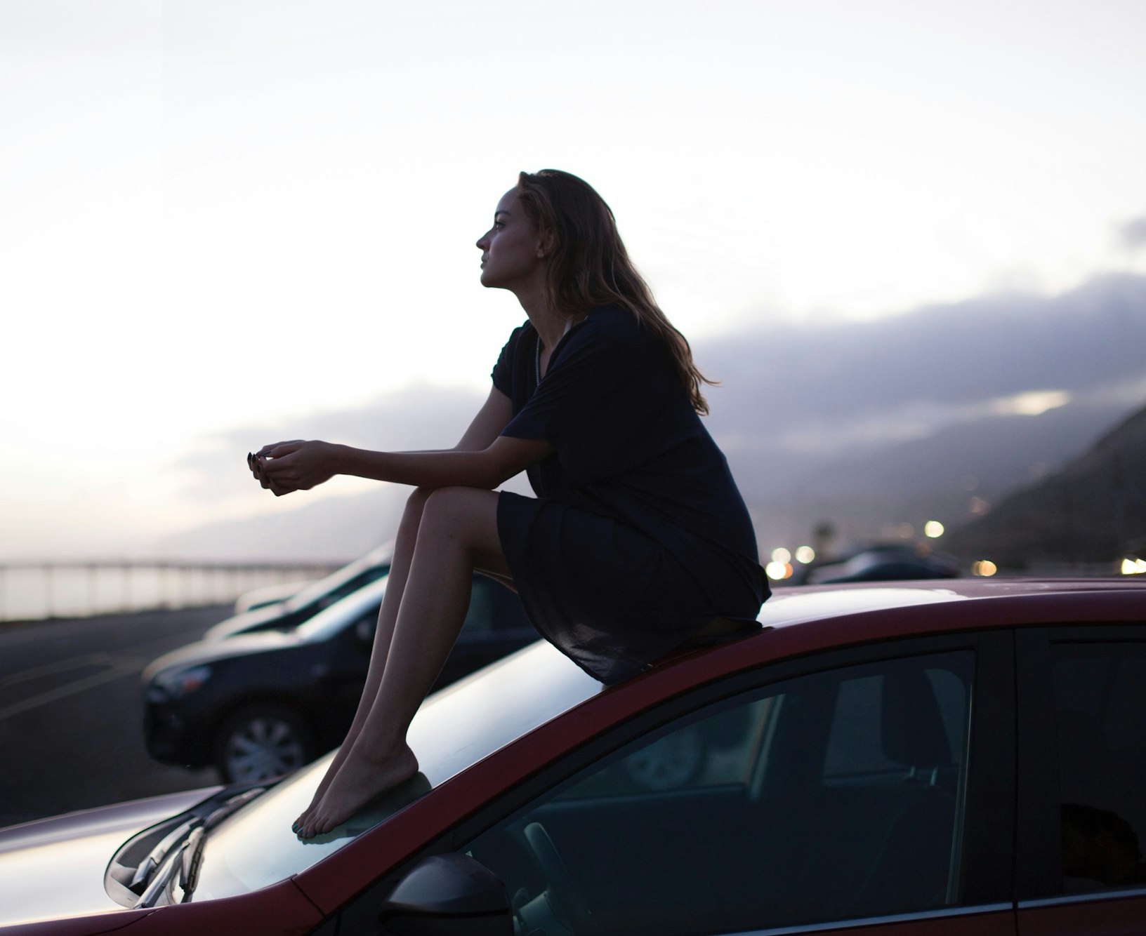 A young woman sits on the hood of a parked car near the ocean at dusk, with a serene expression and soft, diffused lighting.