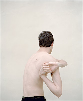 Shirtless man stands against a plain beige background, facing away from the camera, with his arms bent behind his back.