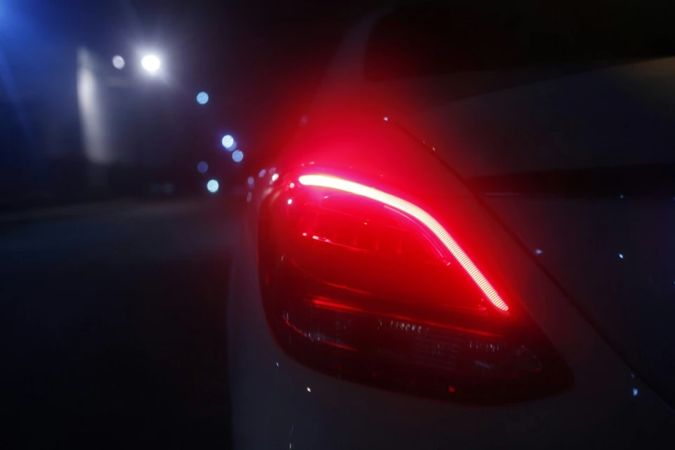 Close-up of white car's rear taillight, illuminated bright red against a dark night backdrop with soft city lights in the distance.