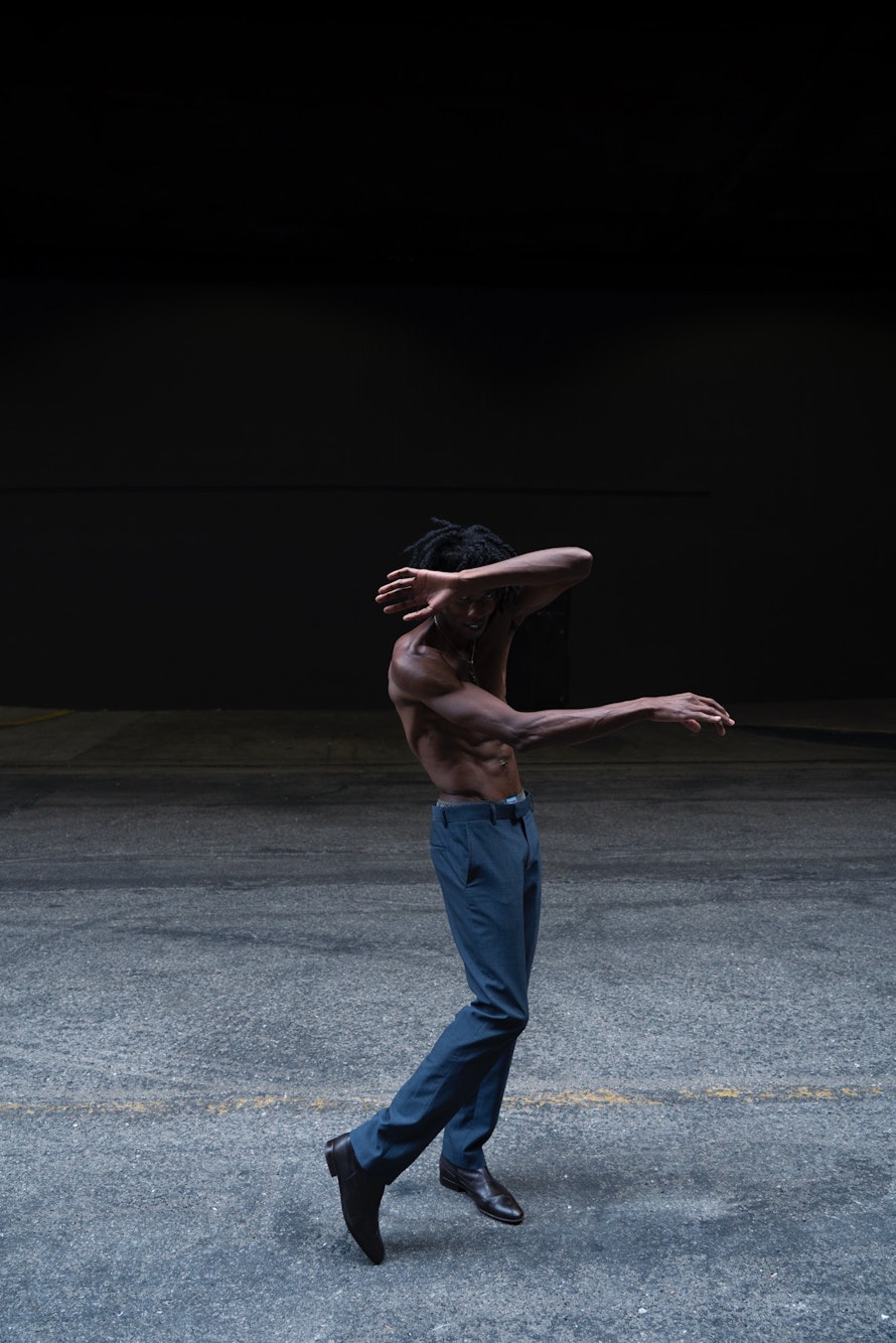 Athletic, shirtless Black man in blue pants dancing on concrete with a dark background. Expressive dance movement in a minimal setting.