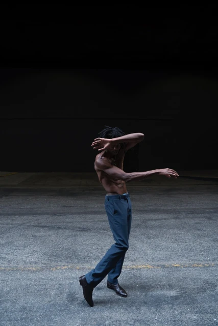 Athletic, shirtless Black man in blue pants dancing on concrete with a dark background. Expressive dance movement in a minimal setting.