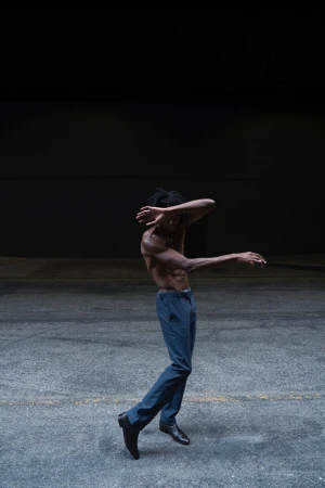 Athletic, shirtless Black man in blue pants dancing on concrete with a dark background. Expressive dance movement in a minimal setting.