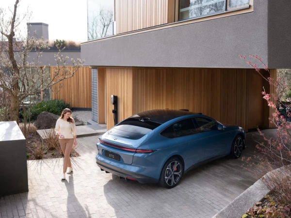 Woman walks near a blue electric car parked in front of a modern house with wood and gray siding, showcasing a contemporary design.