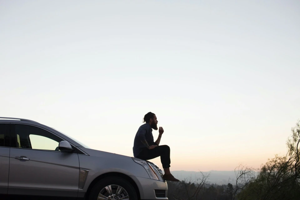 An adult male with a beard sits on the hood of a silver car, with a clear sky background. He is wearing dark pants and a button-down shirt.