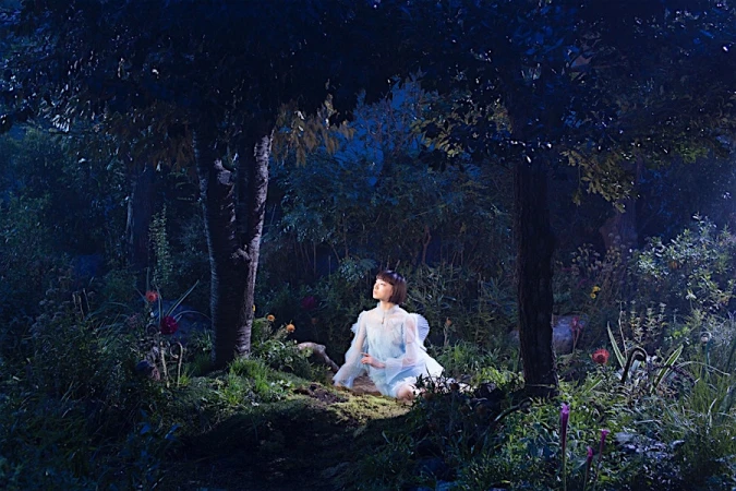 Young woman in a light blue dress seated amidst dense forest foliage at night under dark trees.