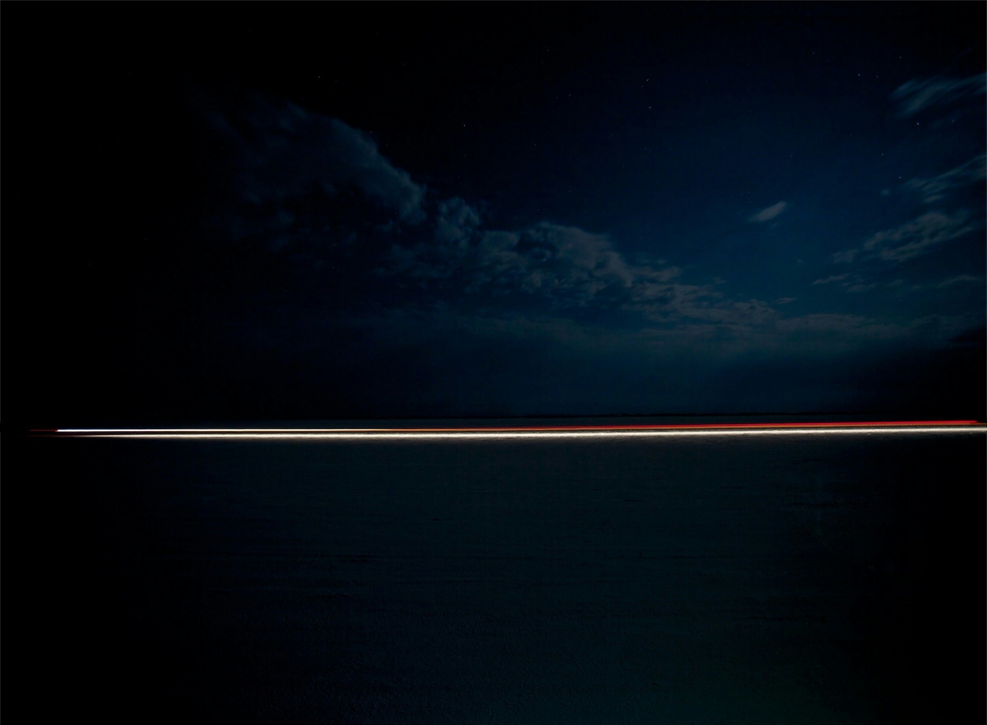Abstract time-lapse photography of light trails across a dark landscape under a cloudy night sky dotted with stars.