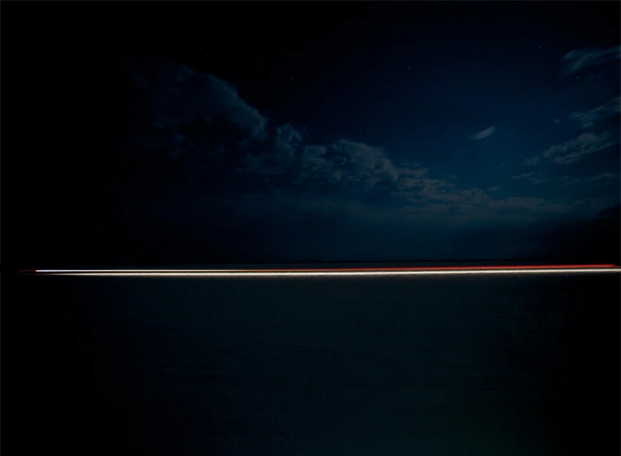 Abstract time-lapse photography of light trails across a dark landscape under a cloudy night sky dotted with stars.