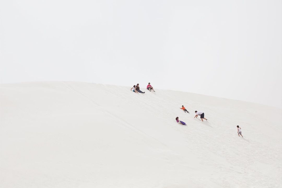 Group of adults and children sledding down a bright sand dune under an overcast sky, conveying a sense of fun and outdoor activity.