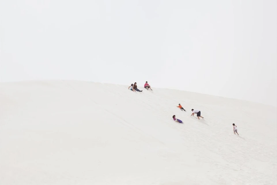 Group of adults and children sledding down a bright sand dune under an overcast sky, conveying a sense of fun and outdoor activity.
