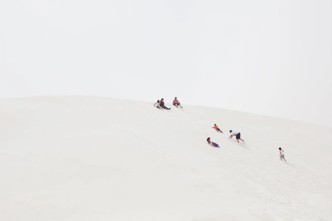Group of adults and children sledding down a bright sand dune under an overcast sky, conveying a sense of fun and outdoor activity.