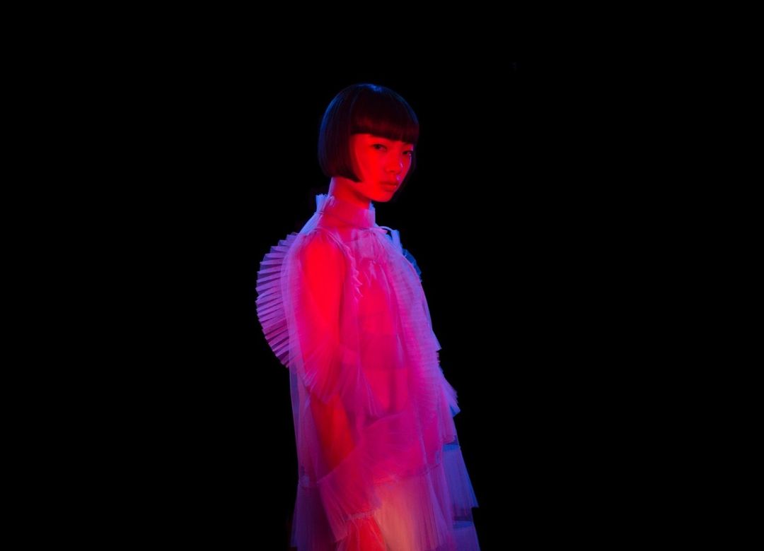 Asian adult female in translucent frilled dress, illuminated by blue and red lights against a black background. Fashion photography.