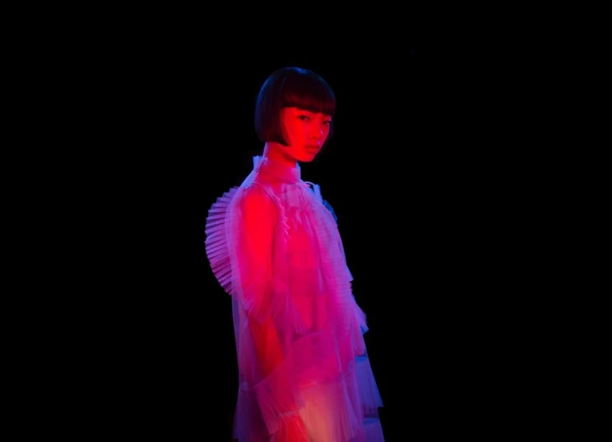 Asian adult female in translucent frilled dress, illuminated by blue and red lights against a black background. Fashion photography.