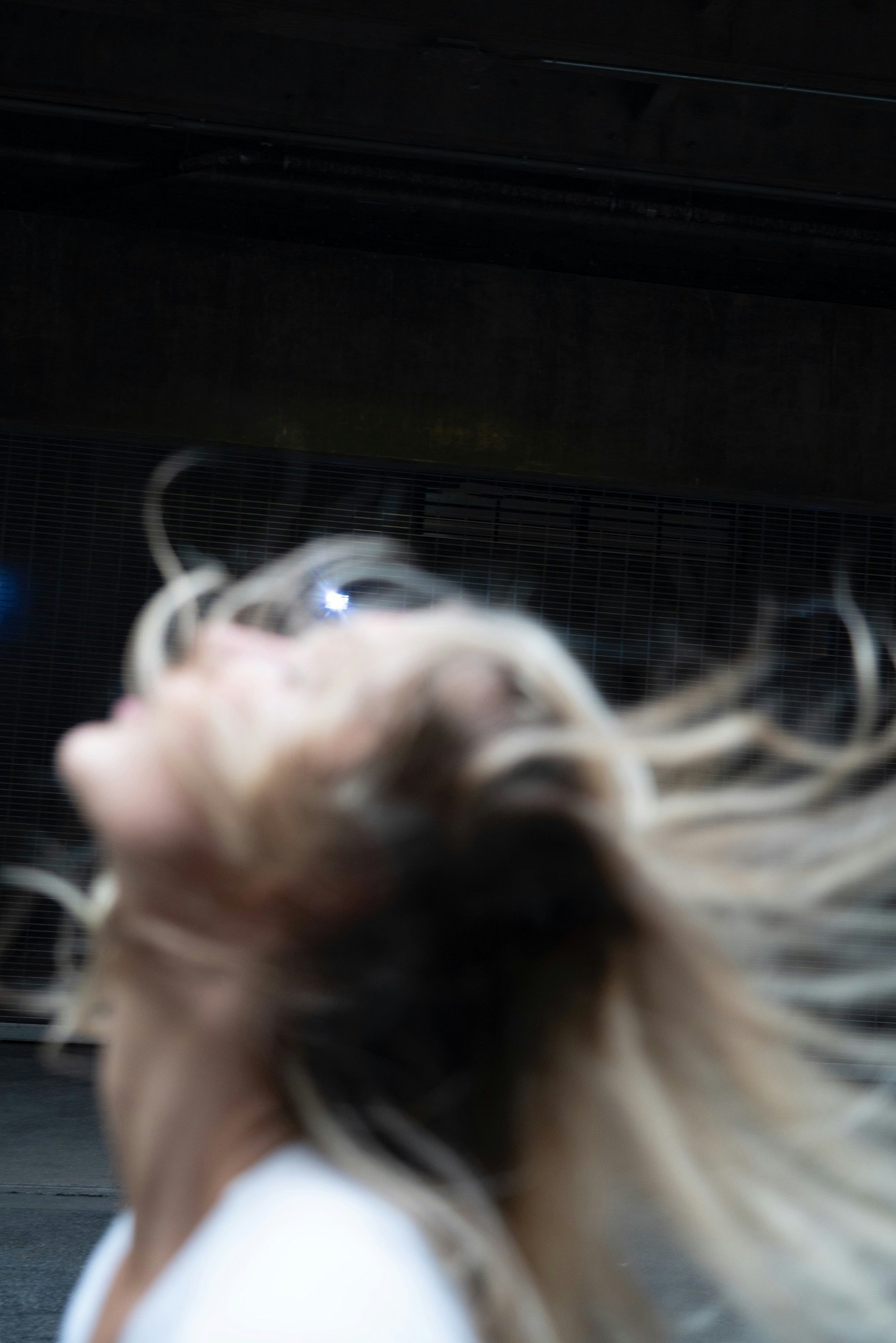 Blurred image of an adult female with long, blonde hair, head tilted back, in an indoor, dark environment.