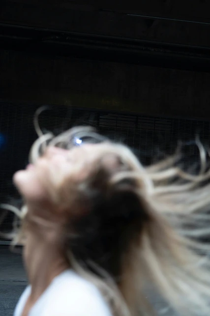 Blurred image of an adult female with long, blonde hair, head tilted back, in an indoor, dark environment.
