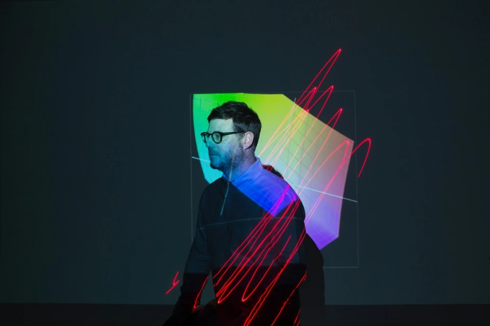 Adult male with glasses stands illuminated by colorful geometric projections and dynamic red light painting in a modern indoor setting.