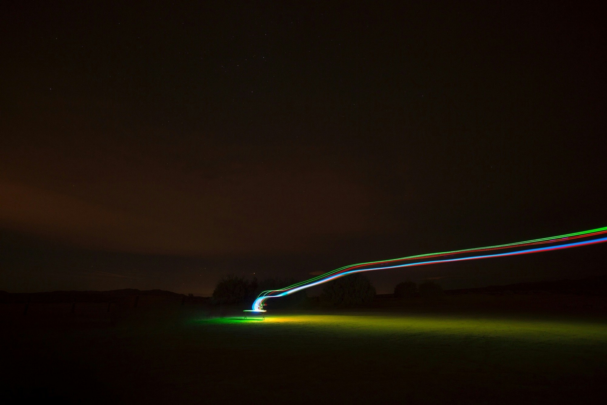 A long exposure captures neon light trails streaking across a dark rural landscape, creating vibrant lines against the night sky.