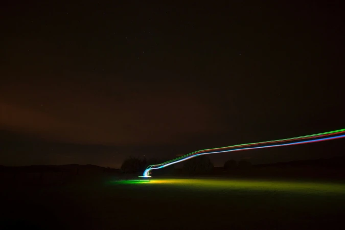 A long exposure captures neon light trails streaking across a dark rural landscape, creating vibrant lines against the night sky.